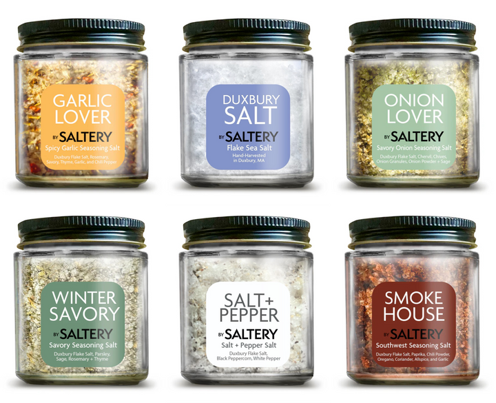 Six glass jars of Saltery seasoning blends arranged in two rows on a white background. The jars feature black lids and labels for Garlic Lover, Dunberry Salt, Onion Lover, Winter Savory, Salt & Pepper, and Smoke House.