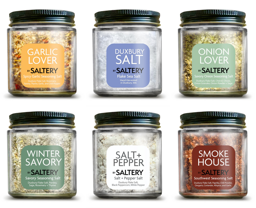 Six glass jars of Saltery seasoning blends arranged in two rows on a white background. The jars feature black lids and labels for Garlic Lover, Dunberry Salt, Onion Lover, Winter Savory, Salt & Pepper, and Smoke House.
