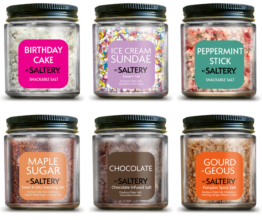 Six clear plastic jars of Saltery snackable salt arranged in a 2x3 grid on a white background. Each jar has a black screw-on lid and a colorful label with a different color and flavor name: Birthday Cake, Ice Cream Sundae, Peppermint Stick, Maple Sugar, Chocolate, and Gourd Geòus.